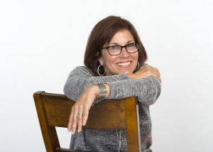 Poet Gianna Russo, photo by Lou Russo. Gianna is sihlouetted on a white background. She has shoulder length, thick, dark hair, dark rimmed glasses and a winning smile. She wears large hoop earrings and a gray sweater. Her arms are draped casually over the back of a chair in front of her.