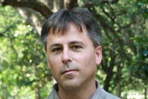 Tennessee poet, Jeff Hardin. You can just see him from the neck up staring at the camera with a straight, close-lipped mouth. He stands under a tree with green leaves for a background.