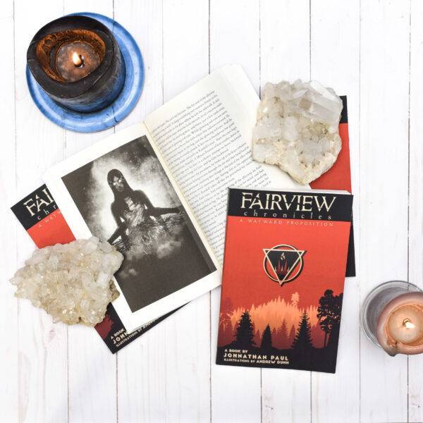 The Fairview Chronicles Vol. 1 arranged on a white washed wood paneling background. Two closed copies prop up a copy which is opened to an image of a woman being disemboweled. Another closed copy and two chunks of quartz hold the book open. Two candles fill negative space on either side of the image.