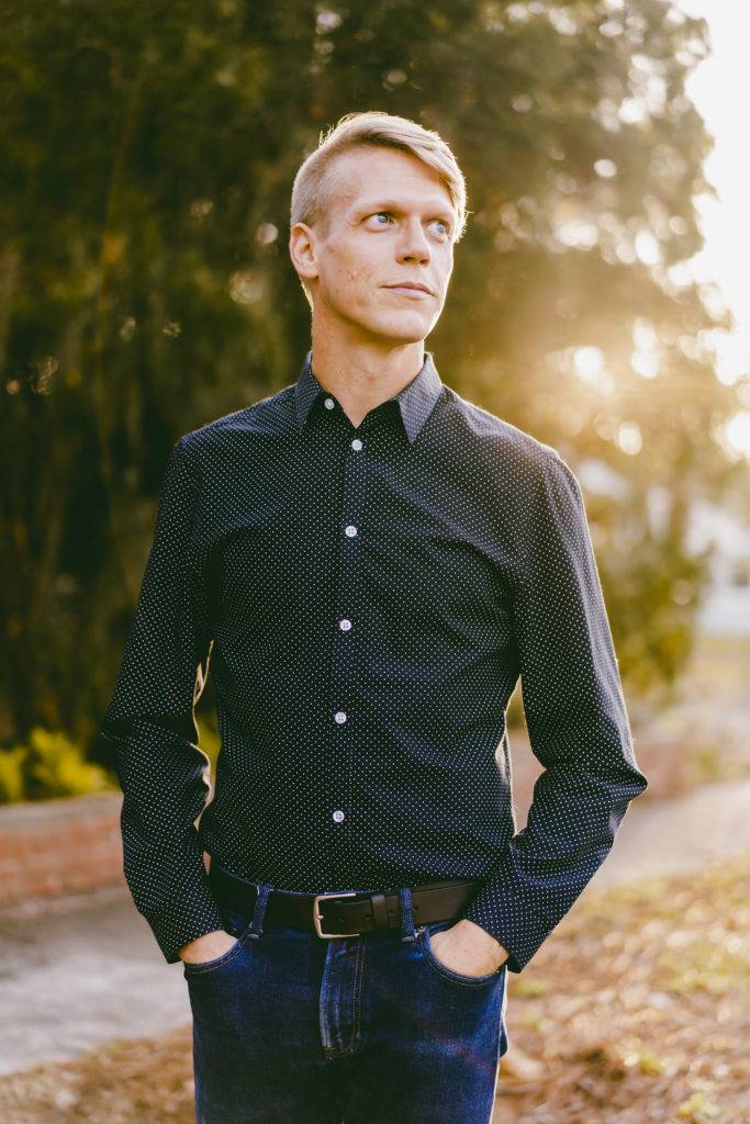Author and journalist, Cooper Levey-Baker. He has blond hair and is standing wearing a dark button-up shirt and jeans. There are trees with sunlight filtering through them in the background.