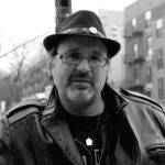 Gerry LaFemina, author of The Pursuit: A Meditation on Happiness. Black and white photo shows LaFemina wearing a fedora and a leather jacket with stout apartment blocks over his left shoulder, and white sky with power poles over his right.