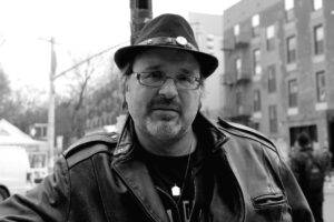 Gerry LaFemina, author of The Pursuit: A Meditation on Happiness. Black and white photo shows LaFemina wearing a fedora and a leather jacket with stout apartment blocks over his left shoulder, and white sky with power poles over his right.