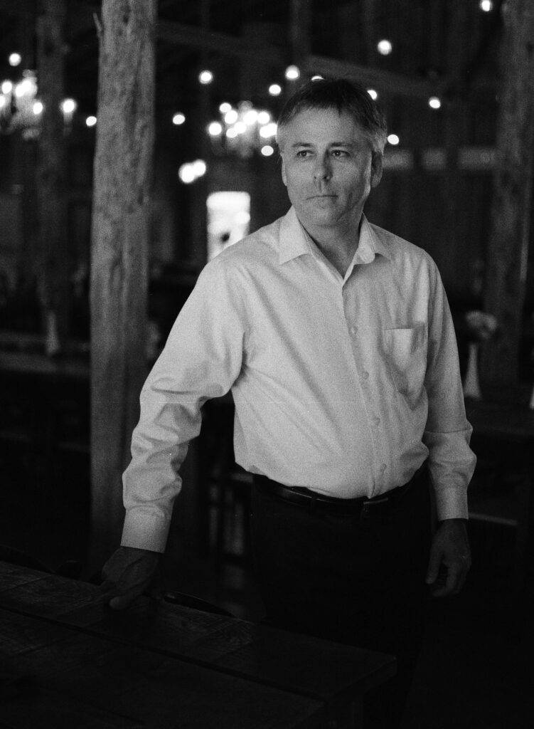 Jeff Hardin, poet, looks pensive in a black and white photo in which he wears a white shirt and black trousers. It's night and strings of lights twinkle behind him.