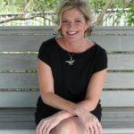 Poet Kate Sweeney with arms and legs crossed and grinning from the porch swing in a little black dress. She has a blond bob haircut and a cheerful smile.