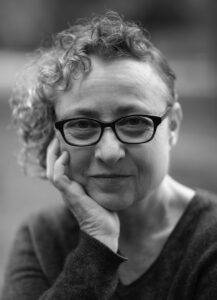 Author Sue Mell, a study in black and white. She rests her chin on one hand, her assymetrical hair lies in waves on the same side. She wears glasses and has just a hint of a smile, or is it a smirk?
