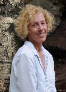 Susanne Davis, author of Gravity Hill. She has chin-length blonde curls, and she is wearing a relaxed cotton button-up shirt. 