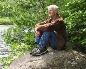 George Drew, sitting on a rock. He has white hair and a heavy brown shirt, jeans, and gym shoes. At his back is a wall of green plants. The light filters onto his face through leaves we cannot see.