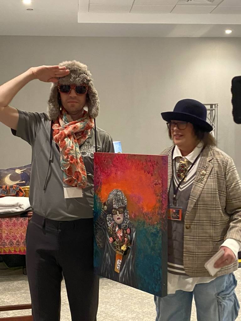Pulpwood Queen's founder Kathy L. Murphy's son-in-law dresses up in a hat, scarf, and glasses and salutes with author Tracy Lea Carnes (The Darlings of Sundance) and a painting of her book cover. Book cover art by Pulpwood Queens founder Kathy L. Murphy.