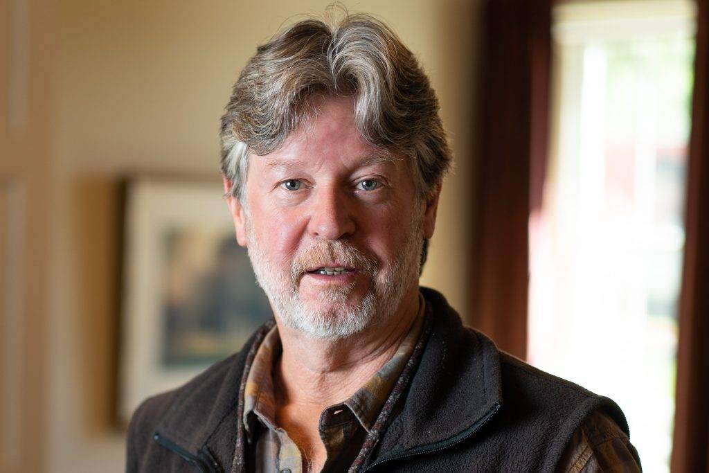 Author, Michael Gills. Photo shows a White man with gray hair and beard wearing a quilted vest and button up shirt with sleeves rolled up--all in brown tones. He has a pair of glasses slung around his neck on a granny cord.