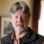 Author, Michael Gills. Photo shows a White man with gray hair and beard wearing a quilted vest and button up shirt with sleeves rolled up--all in brown tones. He has a pair of glasses slung around his neck on a granny cord.