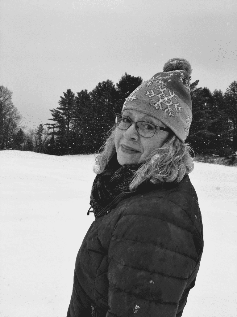 Author Susan O'Dell Underwood. A study in black and white with susan in glasses, shoulder-length blond hair, and a knitted cap. She's looking over her left shoulder at the photographer with snow and trees in the distance.