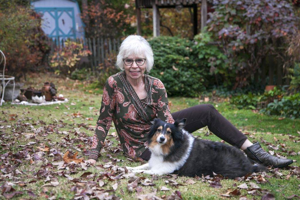 Poet Linda Parsons with her beloved Frankie Bear in her garden. Linda has white, chin length hair, glasses and an inviting smile. Frankie Bear is a tri-colored Sheltie, with his white paws crossed posing for the camera. Autumn leaves surround them.