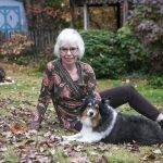Poet Linda Parsons with her beloved Frankie Bear in her garden. Linda has white, chin length hair, glasses and an inviting smile. Frankie Bear is a tri-colored Sheltie, with his white paws crossed posing for the camera. Autumn leaves surround them.