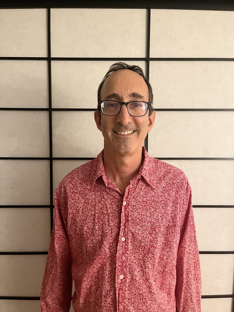 Author David Rothman. David is a light skinned man wearing a red shirt with a white dot pattern. He has short dark hair, a high forehead, black-rimmed glasses, and an engaging smile. He is clean-shaven.