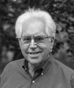 Author Tom Shachtman, with a crown of white hair around a tanned face. The photo is black and white, and he wears a button-down shirt and wire-rimmed glasses. He has a cheeky smile.
