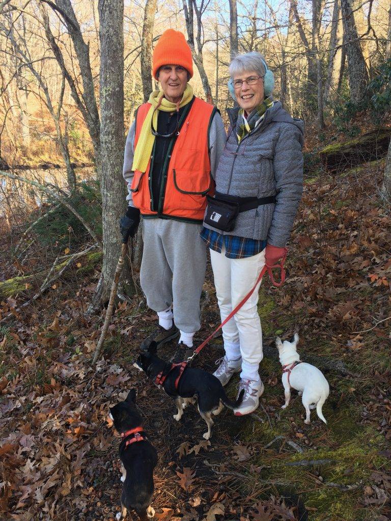 Sam and Vicki Pickering on a wooded hillside with three small dogs on leads. Sam wears bright orange High visibility cap and vest. Vicki wears white jeans and a gray jacket. Both humans are smiling.