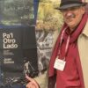 Author Juan Ochoa at #AWP23. Juan is wearing a brown Trilby hat and a red shirt and scarf. He has his hat pulled low, with a cheeky grin beneath a gray moustache. He is pointing with his thumb toward an image of his own book cover.