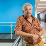 Author Juan Ochoa. Juan is a hispanic man with gray hair and a moustache. He is wearing a short-sleeved button-up shirt. His posture is casual. There is a bright turquoise blue wall behind him.