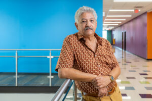 Author Juan Ochoa. Juan is a hispanic man with gray hair and a moustache. He is wearing a short-sleeved button-up shirt. His posture is casual. There is a bright turquoise blue wall behind him.
