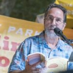 Poet, Neil Shepard, reads from a poetry book. He is in front of a microphone wearing a Hawaiian shirt in blue tones. He has light skin, wire framed glasses, and a gray beard. Behind him is a yellow sign with red and white lettering.