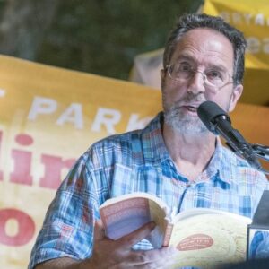 Poet, Neil Shepard, reads from a poetry book. He is in front of a microphone wearing a Hawaiian shirt in blue tones. He has light skin, wire framed glasses, and a gray beard. Behind him is a yellow sign with red and white lettering. 