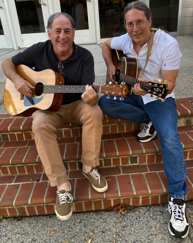 Editors Bob Kunzinger and Drew Lopenzina sitting on the steps of Old Dominion University. Both wear smiles and sneakers and hold guitars, but appear to be laughing too much to play. Bob has thinning gray hair, and drew has a salt and pepper ponytail.