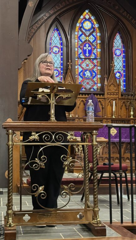 Poet, Darnell Arnoult reading from her new poetry collection, Incantations. Darnell is standing in front of three beautiful stained glass windows, and she looks radiant reading from a gilded lectern. Darnell is a white woman wearing a black blouse. Her hair is shoulder length and streaked with white. She wears glasses.