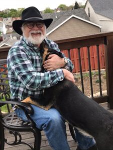 Author Michael Simms playing with his dog. He wears a blue plaid shirt and jeans with cowboy hat, glasses, and a white beard that can't quite hide a wide smile.