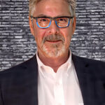 Author Dean Monti. Dean wears a white shirt and black jacket. His collar is open. His gray and white hair and beard are trimmed short, and he looks straight at the camera through a pair of glasses with blue inner frames. Behind him is a gray brick wall.