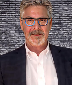 Author Dean Monti. Dean wears a white shirt and black jacket. His collar is open. His gray and white hair and beard are trimmed short, and he looks straight at the camera through a pair of glasses with blue inner frames. Behind him is a gray brick wall.