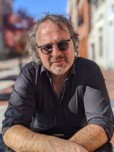 editor, Gerry LaFemina, photo by Mercedes Hettich. Gerry is relaxed, leaning forward on his elbows. He wears sunglasses and a gray button up shirt. His graying hair, thinning on top, is wild, a short salt-and-pepper beard, and an impish smile define Gerry LaFemina in this photo.