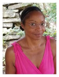 Allison Joseph is a chocolate skinned woman and in this picture she wears a bright pink sleeveless top. she has a gentle close-lipped smile, and wire-rimmed glasses. Her dark hair is pulled back.She sits in a natural setting with an old stone wall behind her.