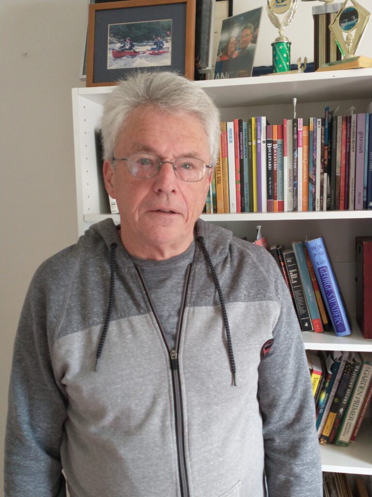 Steve Putnam, The Academy of Reality author. He has nearly all white hair, glasses, light skin and an open expression. He wears a gray hoodie, and stands in front of a book shelf.