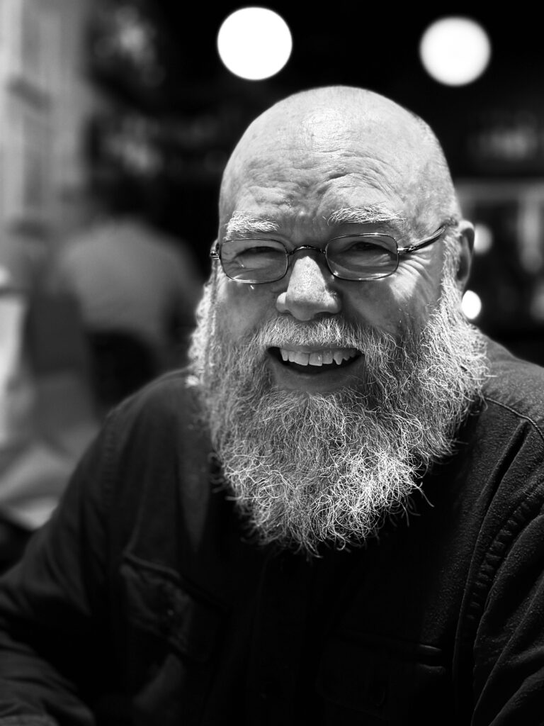 Author Michael Amos Cody. Here we see his head and shoulders. He has a bald head, wire frame glasses, a bushy white beard and moustache, and a big smile. He wears a black T-shirt.