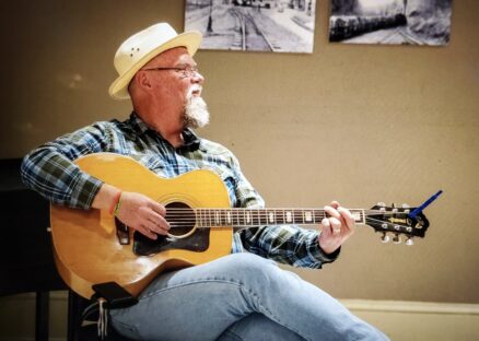 Author, musician Michael Amos Cody wearing a straw hat and strumming a guitar.