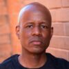 Author Steven T. Moore. Serious face. Steven is a bald Black man with symmetrial features. He wears a black T-shirt and stands in front of a brick wall that harmonizes with the color of his brown skin. His face looks more sad than angry.