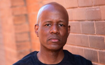 Author Steven T. Moore. Serious face. Steven is a bald Black man with symmetrial features. He wears a black T-shirt and stands in front of a brick wall that harmonizes with the color of his brown skin. His face looks more sad than angry.