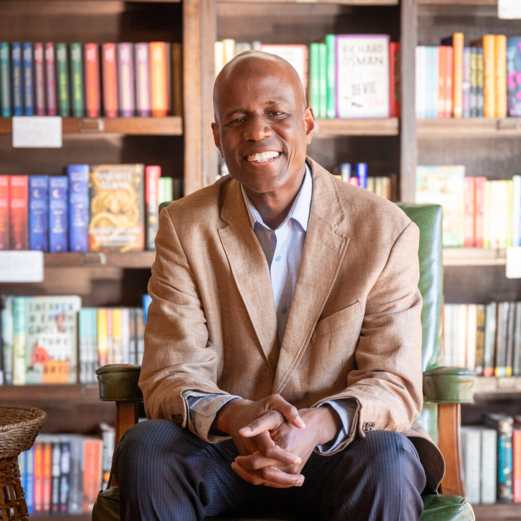 Author Steven T. Moore sits in front of a bookshelf. He wears an open-collared shirt and tan sport coat. His smile is radiant. His skin is the color of milk chocolate. His head is clean-shaven.