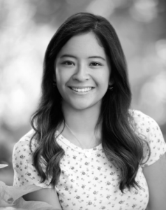 Author Katie Sanyal. Black and white photo shows a young woman with long dark hair and a bright smile. Her blouse has a small print.