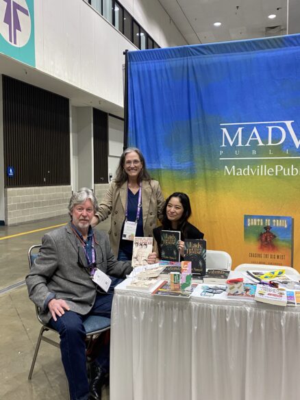Authors Michael Gills and Katie Sanyal with Kim Davis at AWP25 in Los Angeles. They are seated at the booth. Michael has gray hair, Kim has a big grin, and Katie has long dark hair. They hold their books and smile for the camera