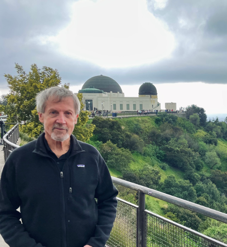 Author Gary Finke. Stands with a dramatic background on a coast with an observatory in the background. Finke has gray hair cut short with a beard on his chin and a wry smile. He wears a dark colored fleece overshirt.