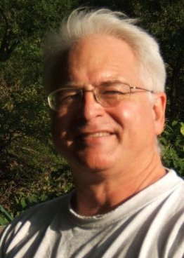 Author Michael Simms. He has white hair, pale skin, wire-rimmed glasses and a warm smile. He wears a white t-shirt and appears to be outside.