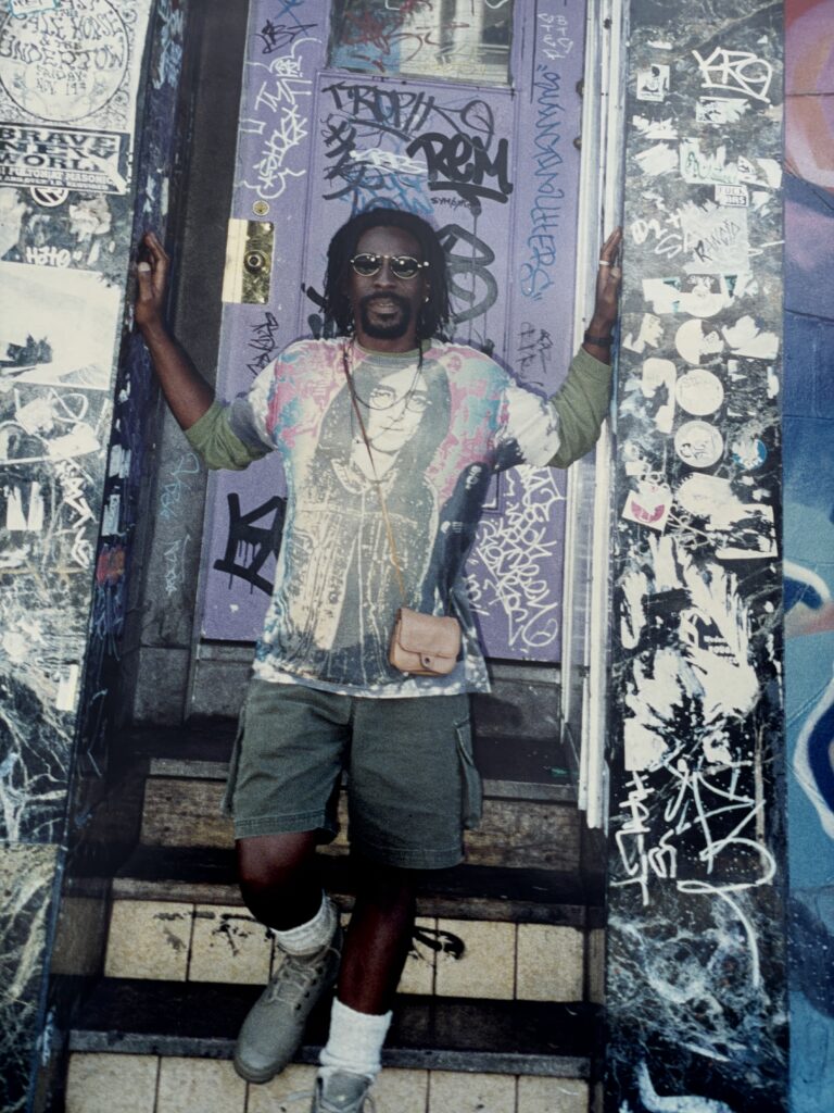 Author Earl S. Braggs with dreads in his hear, dark glasses, and a John Lennon t-shirt. He is posing in a heavily graffitied dorway, and all is in tones of white and blue.