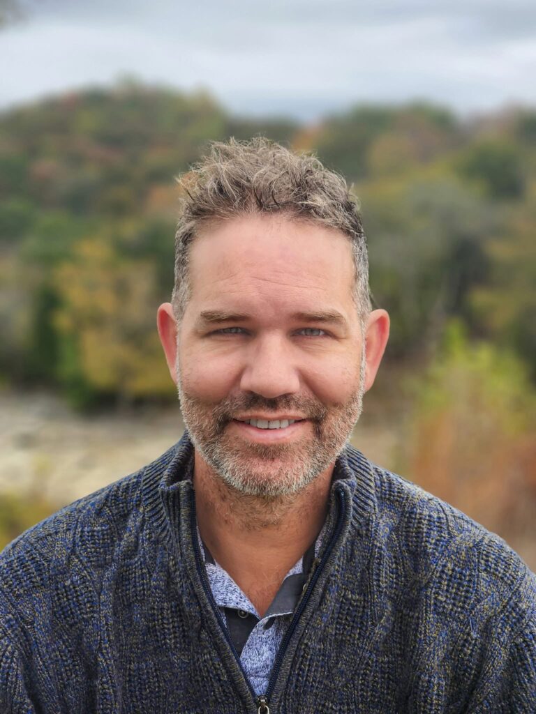 Editor/poet Andrew McFadyen-Ketchum. He is outside with trees burred out behind him. His hair is brown with flecks of gray with short curls on top, and very short sides. He wears a few days' growth of beard and a smile.