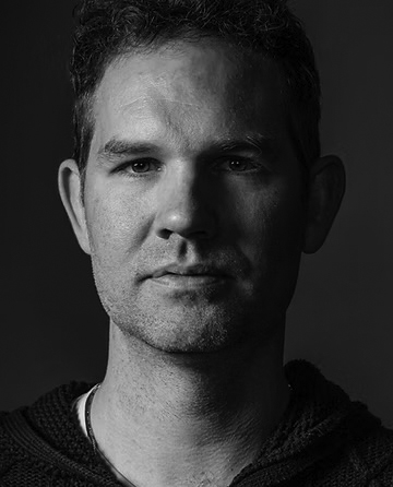Poet and editor, Andrew McFadyen-Ketchum. This black and white photo shows him looking straight ahead without smiling. His face is half in shadow. He is a white man with short hair and a clean-shaven face.