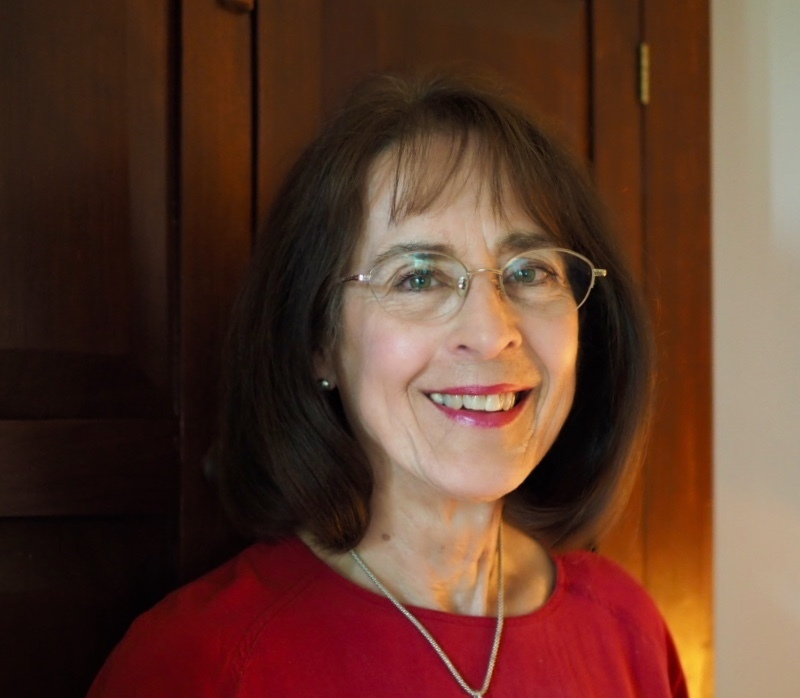 Poet Elaine Fowler Palencia. She is a white woman of indeterminate age with dark hair and glasses. She offers a welcoming smile and wears red lipstick to match her red top.