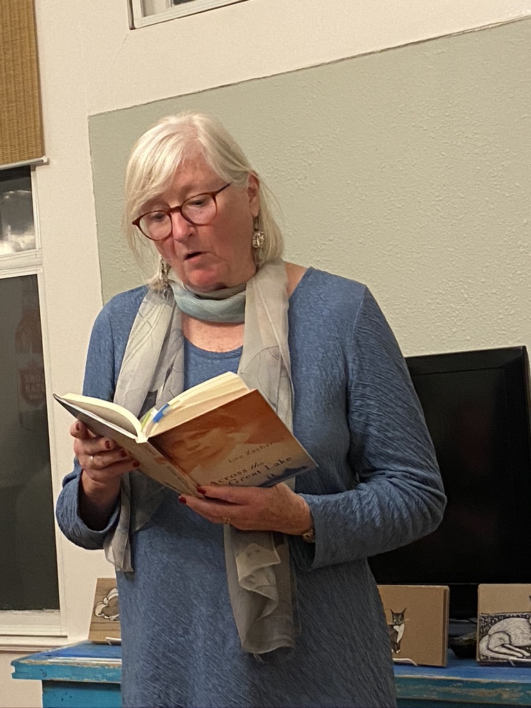 Woman with white hair and a blue blouse with a scarf draped around her neck reads from an orange book.