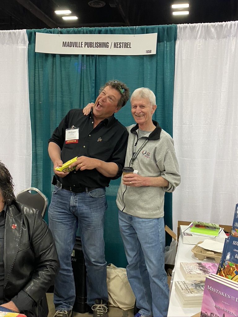 Two men stand in front of a sign that says Madville Publishing / Kestrel