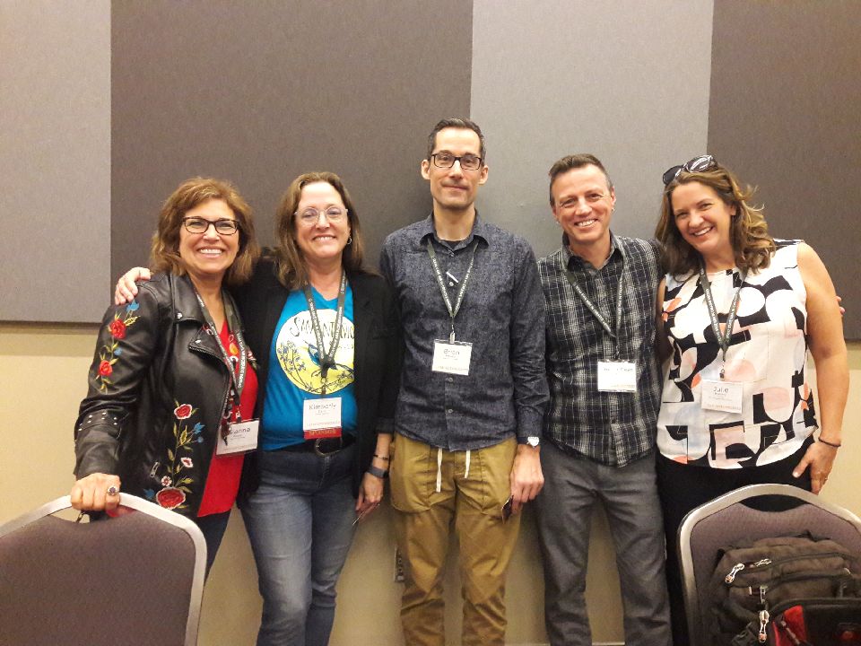 five people posing in front of a tan wall at a convention center. All wear nametags.
