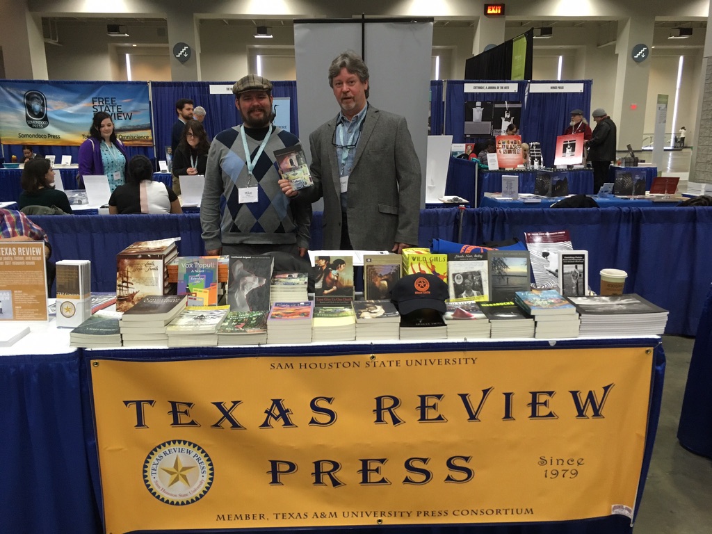 Current Madville authors, Mike Hilbig and Michael Gills stand at TRP's table at AWP 2017. Gills holds his book from that year. There's a bit yellow sign with blue lettering on the front of the table that says Texas Review Press